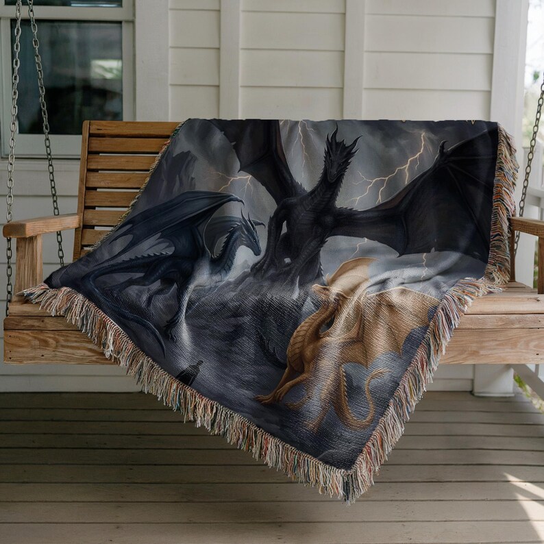 May include: A woven throw blanket featuring a fantasy scene with three dragons in shades of black, gray, and tan. The dragons are set against a stormy sky with lightning. The blanket has a fringed edge and is draped over a wooden swing.
