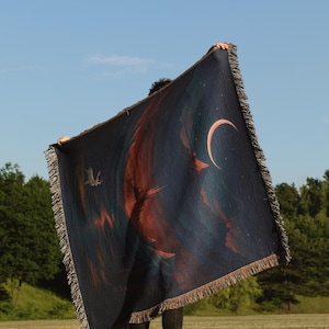 Peut inclure: Une couverture bleu fonc&eacute; avec un croissant de lune et un motif abstrait rouge et orange. La couverture a une bordure &agrave; franges et est tenue contre un ciel bleu et des arbres verts.