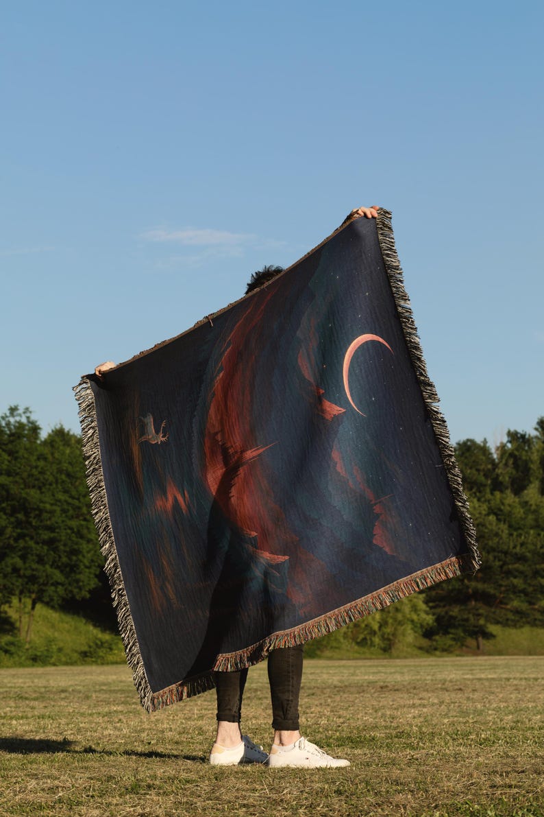 Peut inclure: Une couverture bleu fonc&eacute; avec un croissant de lune et un motif abstrait rouge et orange. La couverture a une bordure &agrave; franges et est tenue contre un ciel bleu et des arbres verts.
