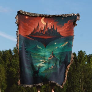 Peut inclure: Une couverture en tapisserie repr&eacute;sentant un paysage fantastique avec un ch&acirc;teau, des montagnes et un cerf. La couverture a une bordure &agrave; franges et une palette de couleurs de bleus profonds, de verts et de rouges. Le ciel est un d&eacute;grad&eacute; de rouge et de bleu.
