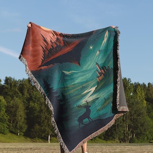 Peut inclure: Une couverture tiss&eacute;e avec un motif de paysage repr&eacute;sentant un cerf, un ch&acirc;teau et une for&ecirc;t. La couverture a une bordure &agrave; franges et une palette de couleurs compos&eacute;e de vert d'eau, de marron et de bleu marine. La couverture est tenue contre un ciel bleu.