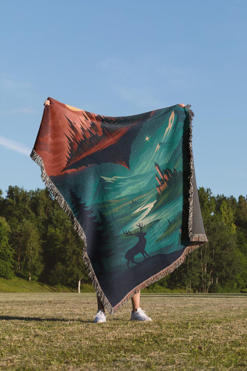 Peut inclure: Une couverture tiss&eacute;e avec un motif de paysage repr&eacute;sentant un cerf, un ch&acirc;teau et une for&ecirc;t. La couverture a une bordure &agrave; franges et une palette de couleurs compos&eacute;e de vert d'eau, de marron et de bleu marine. La couverture est tenue contre un ciel bleu.