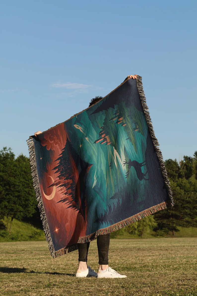 Peut inclure: Une couverture tiss&eacute;e avec une sc&egrave;ne de for&ecirc;t dans les tons de teal, rouge et bleu marine. Le motif comprend des arbres, un croissant de lune et des &eacute;toiles. La couverture a une bordure &agrave; franges et est tenue contre un ciel bleu.