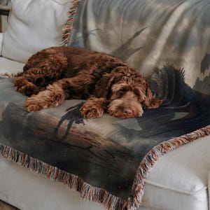 May include: A brown dog rests on a throw blanket draped over a white sofa. The blanket features a fantasy scene with a dragon, a figure, and a dark, misty background. The blanket has a fringed edge.