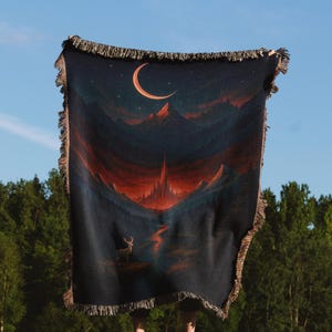 Peut inclure: Une couverture tiss&eacute;e bleu fonc&eacute; repr&eacute;sentant un paysage avec un croissant de lune, des montagnes et un cerf. La couverture a une bordure &agrave; franges et est tenue contre un ciel bleu et des arbres verts.