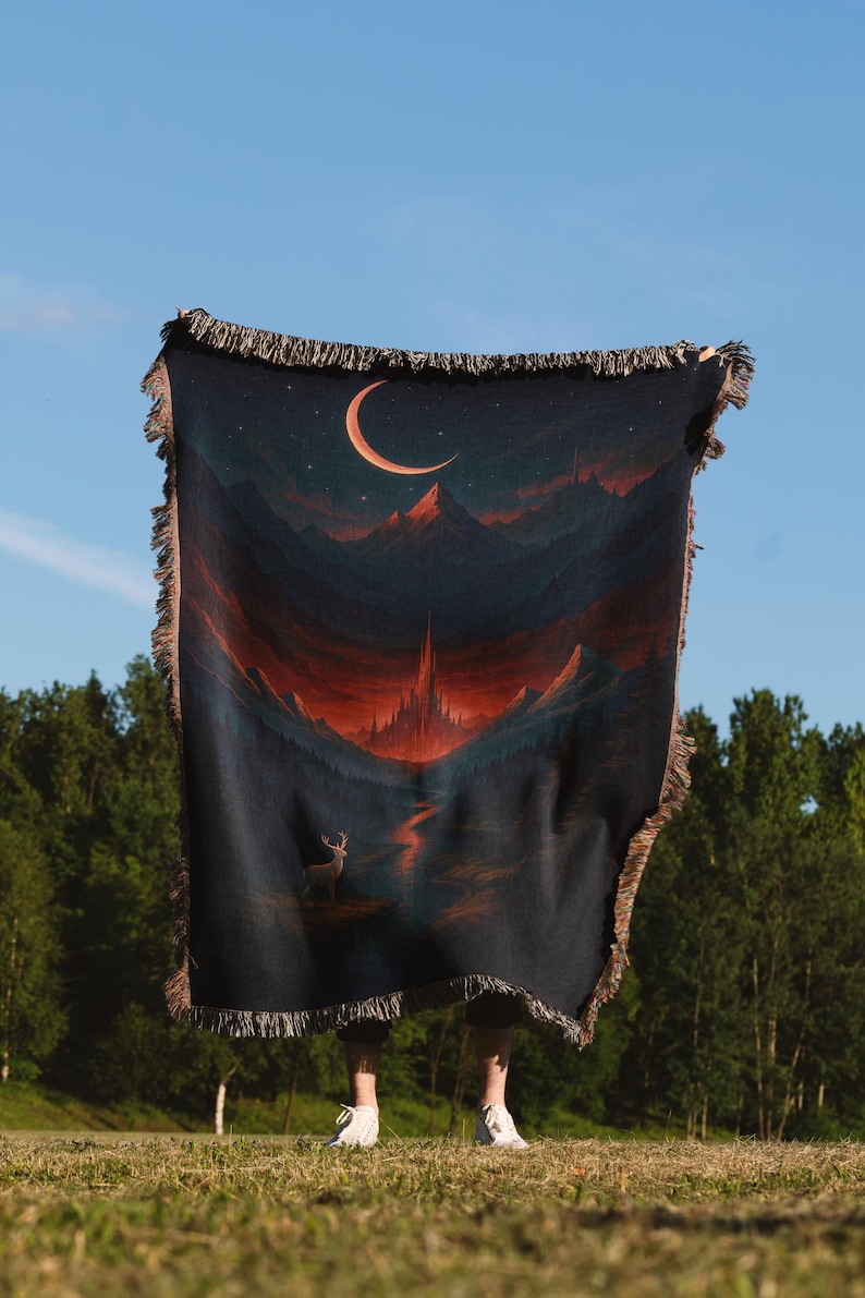 Peut inclure: Une couverture tiss&eacute;e bleu fonc&eacute; repr&eacute;sentant un paysage avec un croissant de lune, des montagnes et un cerf. La couverture a une bordure &agrave; franges et est tenue contre un ciel bleu et des arbres verts.