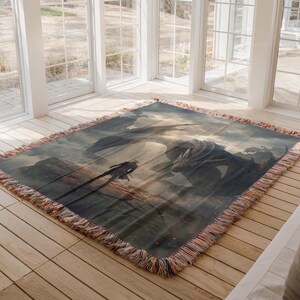 May include: A woven throw blanket featuring a fantasy scene with a dragon and a figure on a mountain. The artwork is in shades of gray, brown, and blue, with a fringed edge.
