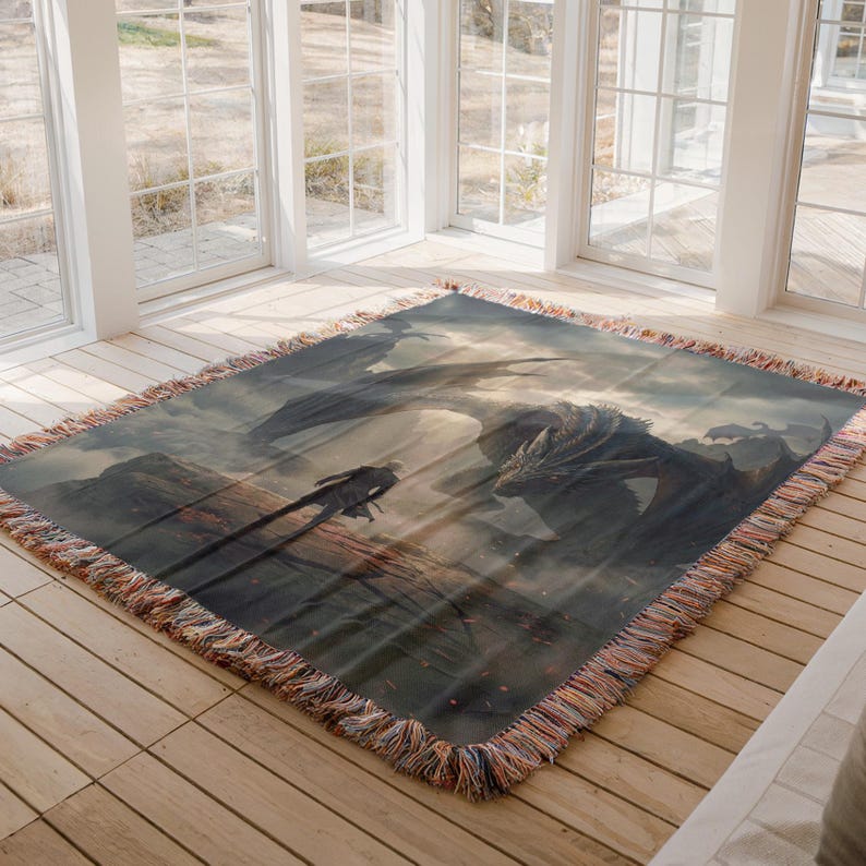 May include: A woven throw blanket featuring a fantasy scene with a dragon and a figure on a mountain. The artwork is in shades of gray, brown, and blue, with a fringed edge.