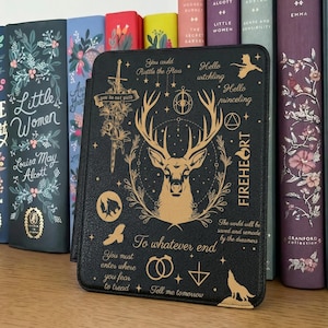 May include: Black book cover with a gold deer design, text that reads "FIREHEART", and other mystical symbols. The cover is leaning against a row of colorful books, including "Little Women" and "Emma".
