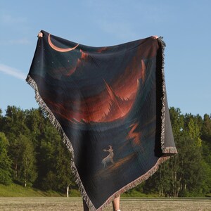 Peut inclure: Une couverture bleu fonc&eacute; avec un motif de paysage repr&eacute;sentant un cerf, un croissant de lune et une cha&icirc;ne de montagnes dans des tons orange et rouge. La couverture a une bordure &agrave; franges.