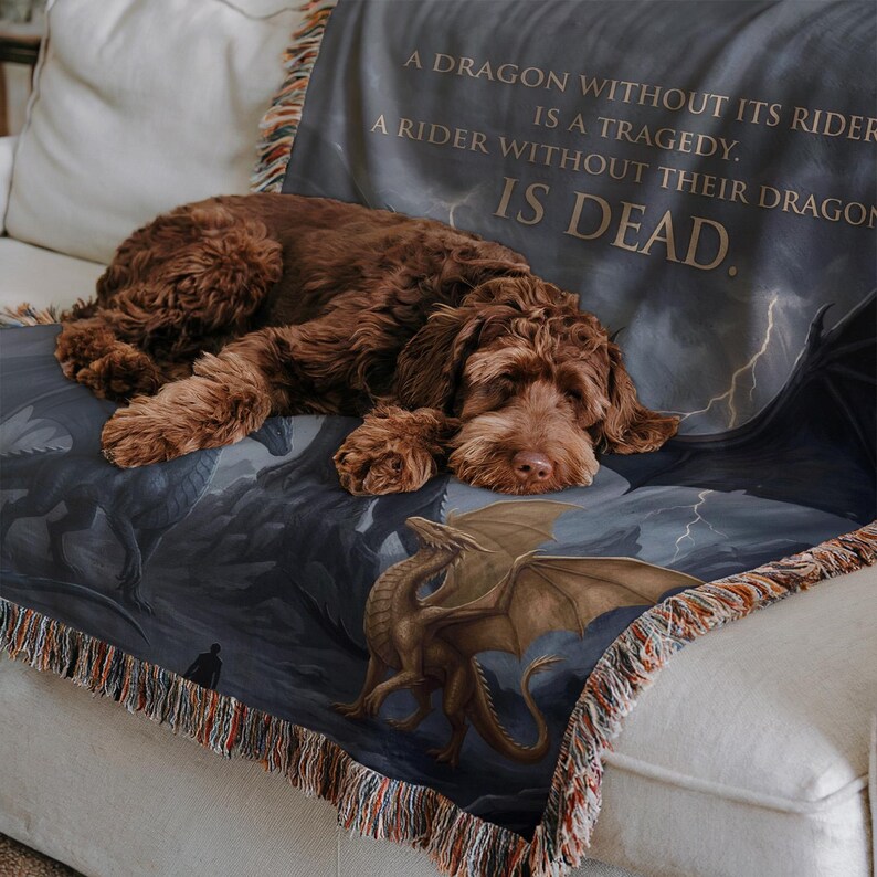 May include: A brown dog rests on a throw blanket with a dragon theme. The blanket features a dragon illustration in shades of brown and gray, with the text "A DRAGON WITHOUT ITS RIDER IS A TRAGEDY. A RIDER WITHOUT THEIR DRAGON IS DEAD."