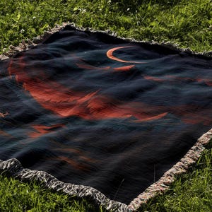 Peut inclure: Une couverture bleu fonc&eacute; avec un motif de croissant de lune et de montagnes dans des tons orange et rouge. La couverture a une bordure &agrave; franges et est pos&eacute;e sur de l'herbe verte.