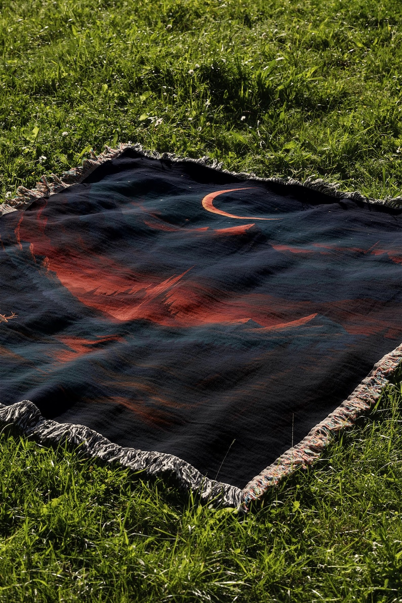 Peut inclure: Une couverture bleu fonc&eacute; avec un motif de croissant de lune et de montagnes dans des tons orange et rouge. La couverture a une bordure &agrave; franges et est pos&eacute;e sur de l'herbe verte.