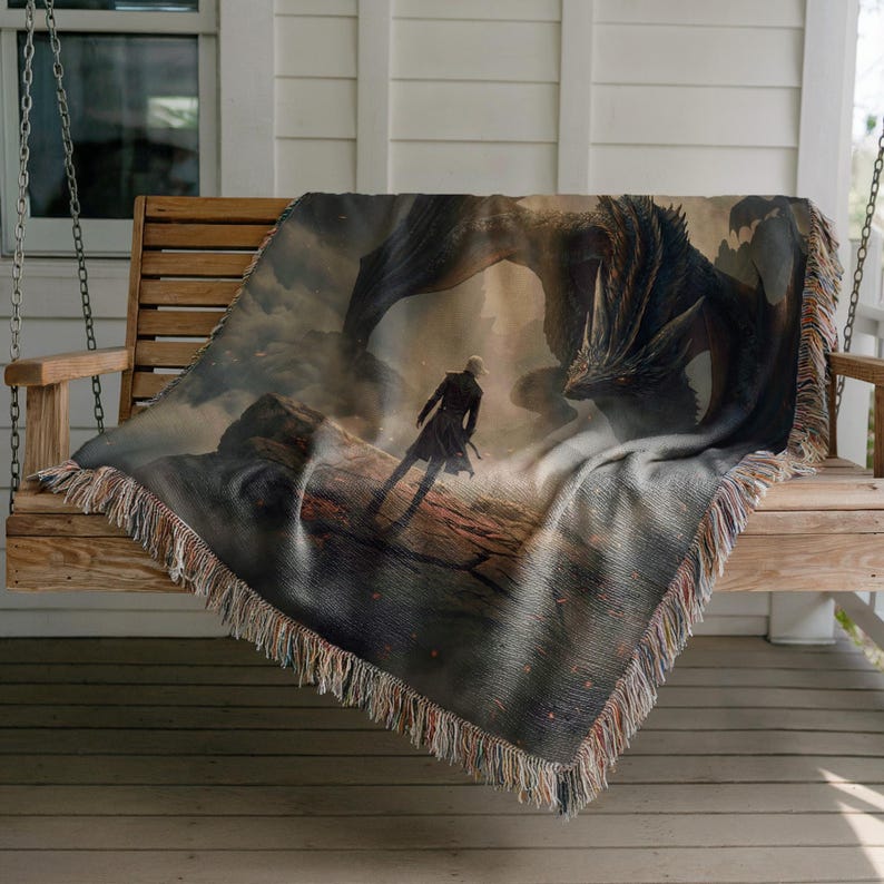May include: A woven throw blanket featuring a fantasy scene with a dragon and a figure in a dark coat. The blanket has a fringed edge and is draped over a wooden swing. The color palette includes shades of gray, brown, and beige.