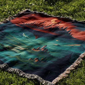 Peut inclure: Une couverture tiss&eacute;e avec un motif de paysage repr&eacute;sentant un ciel nocturne, des montagnes et un croissant de lune. La palette de couleurs comprend des nuances de bleu, de rouge et de vert. La couverture a une bordure &agrave; franges et est pos&eacute;e sur de l'herbe verte.