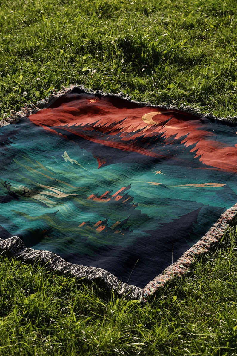 Peut inclure: Une couverture tiss&eacute;e avec un motif de paysage repr&eacute;sentant un ciel nocturne, des montagnes et un croissant de lune. La palette de couleurs comprend des nuances de bleu, de rouge et de vert. La couverture a une bordure &agrave; franges et est pos&eacute;e sur de l'herbe verte.
