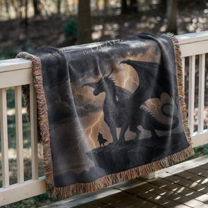 May include: A black throw blanket with a dragon and a figure on a cliff, with the text "A RIDE IS DEAD." The blanket has a fringed edge and is draped over a white railing. The background shows a stormy sky.