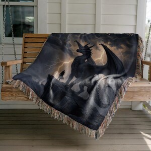 May include: A woven throw blanket featuring a dramatic fantasy scene. A black dragon stands on a rocky peak under a stormy sky with lightning. A figure stands before the dragon. The blanket has a fringed edge.