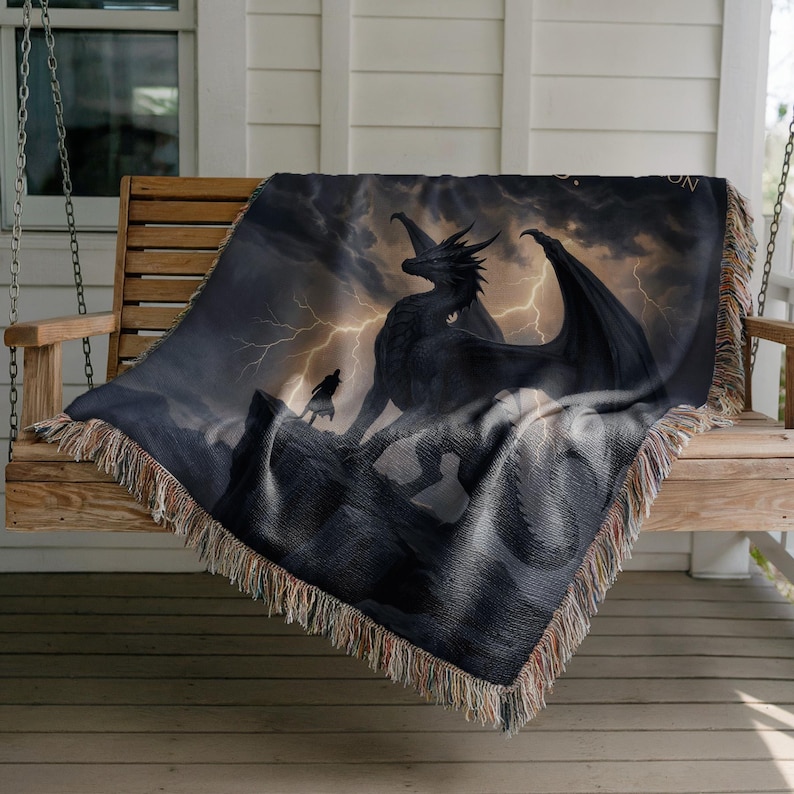 May include: A woven throw blanket featuring a dramatic fantasy scene. A black dragon stands on a rocky peak under a stormy sky with lightning. A figure stands before the dragon. The blanket has a fringed edge.