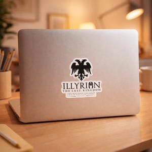 Puede incluir: Una pegatina en un portátil plateado con un emblema de águila bicéfala negra y el texto "ILLYRIAN THE LAST KINGDOM". Debajo hay una línea de texto más pequeña. El portátil está sobre un escritorio de madera.
