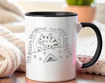 May include: White ceramic coffee mug with a black rim and handle. The mug features a black and white illustration of a cat in bed on a phone with the text "Too sleepy to function too awake to sleep".