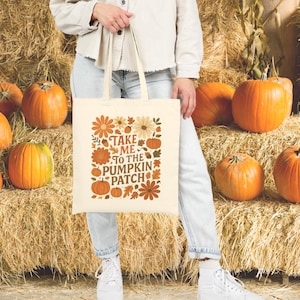 Può includere: Borsa tote color crema con la scritta "Take Me To The Pumpkin Patch" circondata da illustrazioni floreali e di zucche a tema autunnale. La borsa è tenuta davanti a uno sfondo di zucche e balle di fieno.