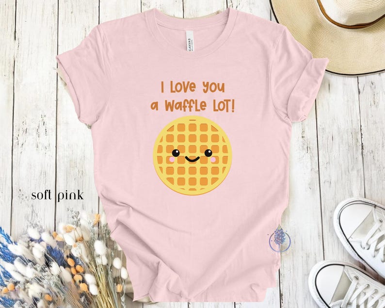 May include: A soft pink t-shirt with the text "I love you a waffle lot!" and a cartoon waffle with a smiling face.