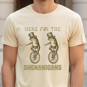 Pode incluir: Uma camiseta bege com o texto "Here for the Shenanigans" estampado na frente. Duas rãs verdes usando chapéus de cartola estão andando de monociclo de cada lado do texto.
