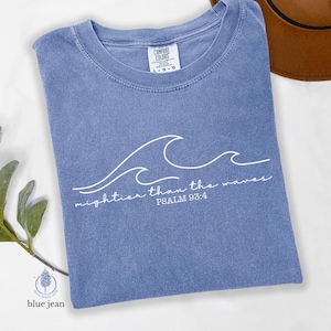 Può includere: Una maglietta Comfort Colors blu jeans con un disegno di onde bianche e la scritta "mightier than the waves PSALM 93:4". La maglietta è piegata e ha un'etichetta con L-G-G.