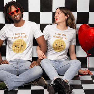 May include: Two people wearing white t-shirts with a waffle graphic and the text "I love you a waffle lot!"  They are sitting in front of a black and white checkered background. A red heart-shaped balloon is in the background.