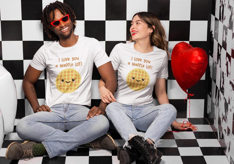 May include: Two people wearing white t-shirts with a waffle graphic and the text "I love you a waffle lot!"  They are sitting in front of a black and white checkered background. A red heart-shaped balloon is in the background.