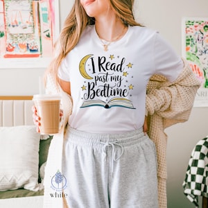 May include: White t-shirt featuring the text "I Read past my Bedtime" in black script, a yellow crescent moon, and yellow stars. The design includes an open book illustration. The shirt is worn with grey sweatpants and a beige cardigan.