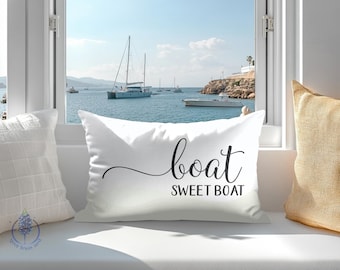 May include: White cushion with the words "boat SWEET BOAT" in black script. The cushion is placed on a white windowsill, with a view of boats on the sea and a blue sky.