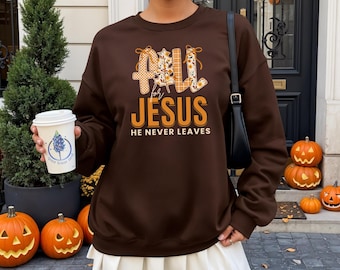 May include: A brown sweatshirt with the text "Fall for Jesus He Never Leaves" in orange and white. The design includes fall-themed elements. The sweatshirt is paired with a white pleated skirt and a black shoulder bag. Pumpkins and a small evergreen tree are in the background.