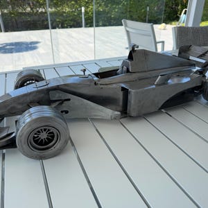 Peut inclure: Une sculpture de voiture de course de Formule 1 grise métallique. La voiture présente des roues détaillées, un design aérodynamique élégant et un aileron arrière. La sculpture est exposée sur une table à lattes blanches, mettant en valeur sa forme et son savoir-faire.