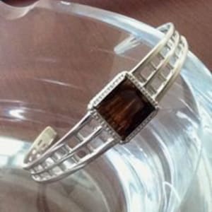 May include: A silver bangle bracelet with a square, brown gemstone set in a silver frame. The bracelet has a geometric design with three horizontal bars.