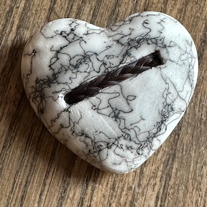 May include: A white heart-shaped ceramic ornament with black lines and a braided hair strand running through a slot in the center.