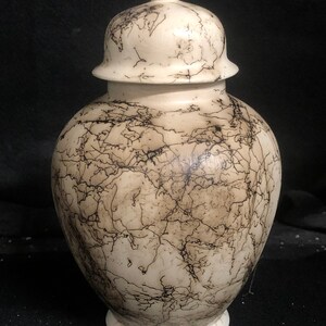 Puede incluir: Un jarrón de cerámica blanca con tapa, decorado con un patrón de craquelado negro. El jarrón tiene un cuerpo redondo y una tapa abovedada.