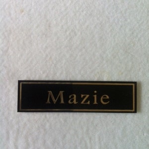 May include: A black rectangular name tag with gold lettering that reads "Mazie".
