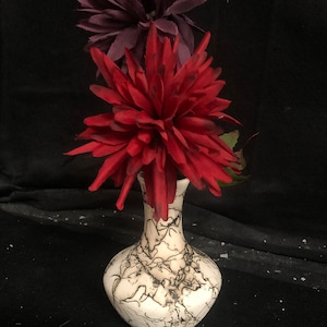 May include: A white ceramic vase with a crackled glaze pattern in black. The vase is filled with two artificial flowers, one red and one purple.