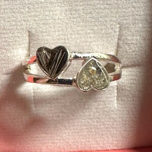 May include: A silver ring with two heart-shaped settings. One heart is filled with dark, textured material, and the other with a lighter, speckled substance. The ring has a double-band design.