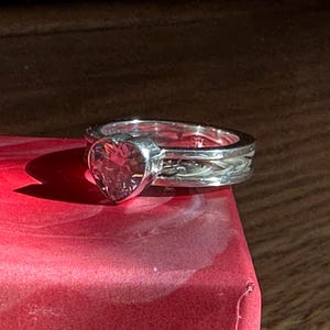 May include: A silver ring with a heart-shaped clear stone set in the centre. The band is braided and has a textured finish.