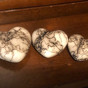 May include: Three heart-shaped ornaments. Each heart is white with a distinctive pattern of black, vein-like lines. The hearts are of different sizes and are arranged on a dark wooden surface. The objects appear to be made of stone or a similar material.
