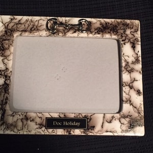 May include: A white ceramic picture frame with a black and brown marbled design. The frame has a silver metal accent at the top and the words "Doc Holiday" are printed on the bottom.