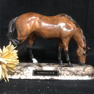 May include: A brown and black horse figurine with a white hoof, standing on a white and black marble base with the inscription "In Memory of FJ".