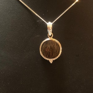 May include: A silver necklace with a round pendant containing a lock of hair. The pendant is surrounded by small clear stones.