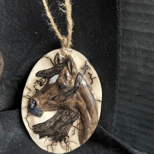 May include: A round, white ceramic ornament with a brown horse head sculpted in relief. The ornament has a brown twine cord for hanging and the text "833" written on the side.