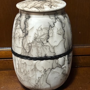 May include: A white ceramic jar with a lid, decorated with black swirling lines. The jar has a black leather band around the middle.
