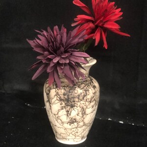May include: A white ceramic vase with a black swirl design, holding two artificial flowers. One flower is red and the other is purple.