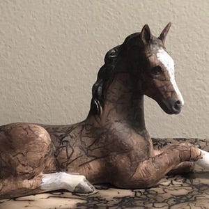 May include: A brown and white ceramic horse figurine in a resting pose. The horse has a white blaze on its face and white markings on its legs. The figurine is detailed with dark brown markings and sits on a textured base.